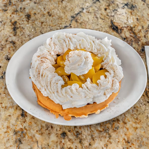 Golden waffle topped with whipped cream and peaches, dusted with cinnamon, on a white plate.
