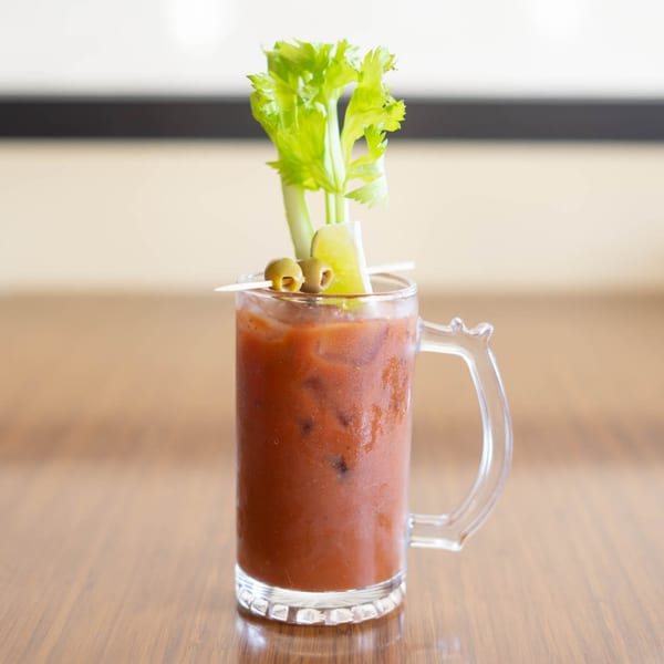 A Bloody Mary cocktail in a mug, garnished with celery, lime, and olives, on a wooden table.