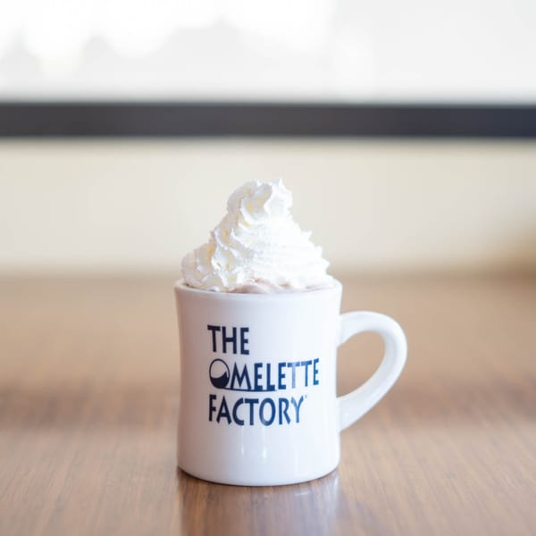 A white Omelette Factory mug filled with hot chocolate and topped with whipped cream.