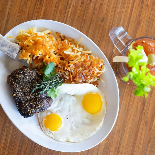 A breakfast plate with steak, two fried eggs, hash browns, and a Bloody Mary on a wooden table.